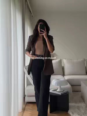 A woman in a blazer and black pants takes a mirror selfie in a well-lit room. She is partially obscuring her face with her phone. The background includes a sofa and curtains, giving a clean, modern aesthetic.