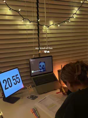 The image depicts a cozy study scene. A laptop is open, displaying a cartoon panda character. A digital clock displays the time 20:55 on a tablet. The scene is illuminated by a string of fairy lights. The foreground shows a person, whose back is to the viewer, writing on a notebook.