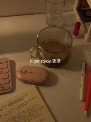 A top-down shot showcases a study desk setting with a warm, inviting ambiance. The composition is dominated by a clear glass mug filled with a light brown beverage, possibly coffee or tea, and rests on a wooden coaster. A pink mouse, a pink keyboard, and a sheet of handwritten notes are arranged on the surface. A glass of water and a red pen alongside a white pen are placed to the right of the mug. The lighting suggests a cozy, nighttime or indoor setting, emphasized by the presence of the mug. The overall color palette is soft and warm, with browns, pinks, and beige dominating.