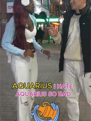 A street interview scene at night. A woman with long burgundy hair, wearing a white top with a light-blue jacket and white pants, is holding a microphone and talking to a man who is also holding a microphone. The hook text states 'Aquarius I hate Aquarius so bad'. An Aquarius sign is positioned at the bottom of the image, with a night city background.