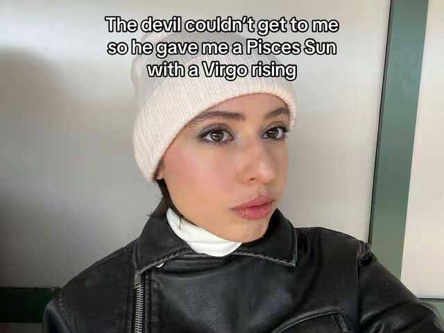 A selfie-style shot of a woman wearing a white beanie, a black leather jacket, and a white turtleneck. The background shows a soft, neutral wall and a green panel. The woman is looking at the camera with a subtle expression and makeup. Text is overlaid at the top.