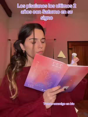 A woman is seated, holding an open book with colorful abstract designs against a soft-focus backdrop. The image is warm, with a focus on the book and the woman's expression. Overlay text in the top left corner in a purple box reads 'Los piscianos los ultimos 2 años con Saturno en su signo', describing the subject as Pisces with the planet Saturn in their sign.
