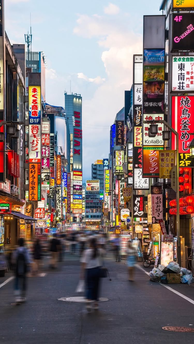 A long vertical shot of a busy street in Tokyo at dusk, with numerous brightly lit signs in Japanese and English. People are blurred as they walk along the street. The sky is a mix of blue and clouds, casting a soft glow over the scene.