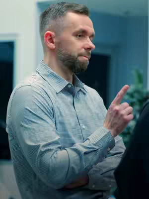 A medium shot of a man with short brown hair and a beard, wearing a button-down shirt, he is pointing his finger upwards, with the background out of focus.