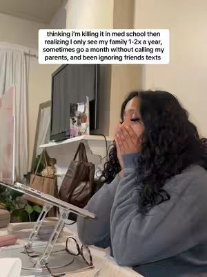 A young woman covers her mouth with both hands in a gesture of emotional distress. She has long, dark, curly hair and is wearing a gray sweater. The background shows a room with a laptop stand holding a laptop, glasses, and other items on a table. A text overlay is at the top of the frame.