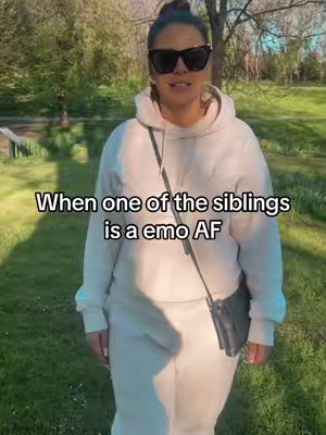 The image shows a woman in sunglasses and a white hoodie, standing on green grass with trees and trees in the background. Text overlayed on the image reads, "When one of the siblings is a emo AF"