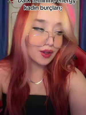 A close-up shot of a woman with long, reddish-pink hair and large-framed glasses. She is looking at the camera with a slight smile and pink lipstick. Text overlayed on the image states 'Dark feminine energy kadın burçları'. The background is blurred and appears to be a room with soft lighting.
