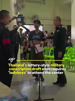 A photograph of a Thai military conscription event. The scene is indoors, with bright lighting and a crowd of people. In the center, a person with short hair and a cropped top stands facing the camera while holding a paper; they appear to be the focus. Uniformed military officers stand nearby, one holding a container and another gesturing. Green plastic chairs are visible in the background. Text overlays at the bottom provide context about the conscription draft and the inclusion of transgender individuals. The overall impression is journalistic or documentary-style.