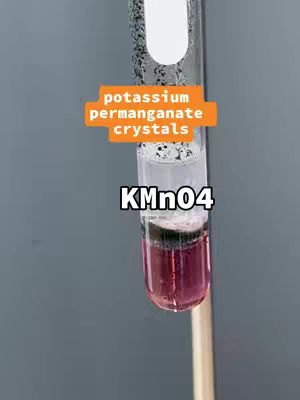 A test tube is filled with a layered solution in a chemistry experiment. The bottom layer is a deep magenta color, while the top layer is colorless, with some dark particles suspended within. The text overlays indicate "potassium permanganate crystals" and "KMnO4". The background is plain gray.