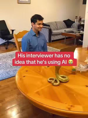A man in a blue shirt sits facing a wooden table. A red banner across the image reads 'His interviewer has no idea that he's using AI'. A background with a chair, rug, and a couch with a table in the background.