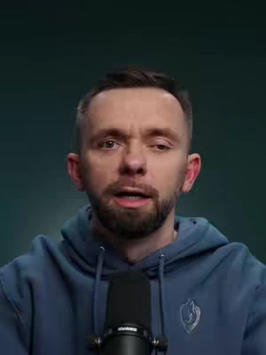 A man with short, dark hair wearing a blue hoodie is centered in a medium close-up. A black microphone is positioned close to his mouth. The background is a solid, muted teal color.