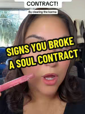 A selfie-style video cover shows a woman with brown hair looking towards the camera with slightly opened lips. She has rosy cheeks and is holding a pink lip gloss in her left hand. The overlay states in bold, yellow text: 'SIGNS YOU BROKE A SOUL CONTRACT' with more text above 'CONTRACT!' and text below 'By clearing the karma'.