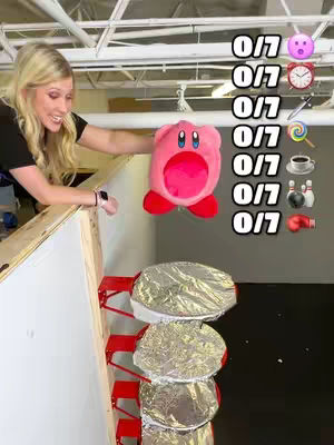 A woman stands in front of a staircase-like structure, holding a pink Kirby plushie. Above a series of platforms, there are numbered indicators with emojis. The scene is set in a nondescript industrial space, and the woman is smiling, with a playful expression that matches the theme of the video.