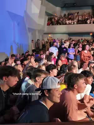 A live scene inside a large auditorium, packed with people. The lighting is bright, and many people are clearly visible in the crowd, creating a sense of a shared experience.