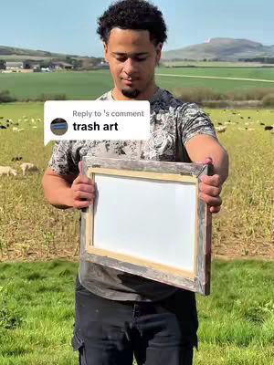 A young man in an olive green patterned t-shirt and black jeans stands outdoors on a sunny day, holding up a framed canvas with a white surface. Behind him is a green field with a few sheep grazing and a rolling green landscape in the distance under a clear blue sky. A comment box with text overlays the image.