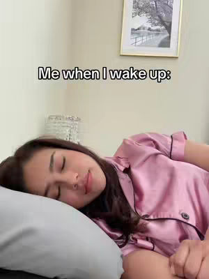 The cover image shows a woman lying in bed, seemingly asleep or just waking up. She's on her side with her eyes closed. The background includes a simple wall, a framed picture, and a decorative lamp. The color palette is soft, dominated by muted tones of pink and white, creating a calm and peaceful atmosphere, with the woman wearing a pink silk pajama. The text overlay reads, "Me when I wake up", placed centrally at the top.