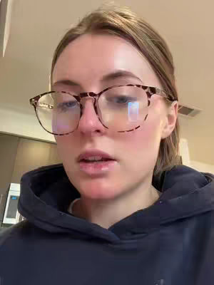 A close-up shot of a woman wearing glasses and a dark hoodie. She is gazing at the camera with a somewhat serious expression. The background is slightly blurred, with hints of a kitchen or living space.