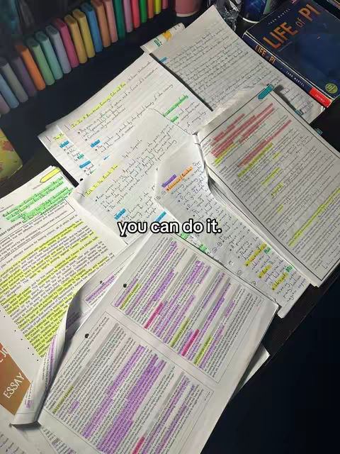 The image shows an overhead view of a desk covered with open books, handwritten notes, and highlighted text. The composition is dominated by the papers, filled with various colors of highlighters, along with a closed book in the top right corner and colorful stationery in the top left, setting a study environment.