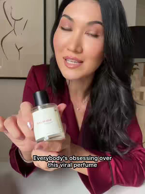 A woman with long dark hair is holding a perfume bottle. She is wearing a burgundy colored robe. The background consists of a wall with an abstract art print in the background.