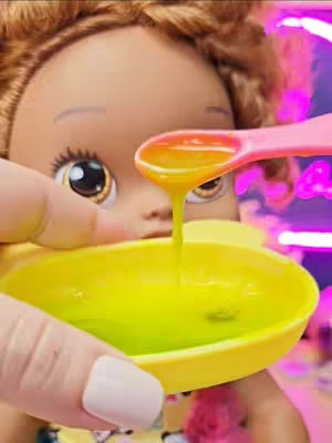 The cover image shows a close-up shot of a Baby Alive doll's face. A hand holds a pink spoon with a yellow liquid over a yellow bowl, from which the liquid is dripping. The doll's eye and part of its face are visible. The background consists of colors like yellow and pink, suggesting a playful or colorful setting.