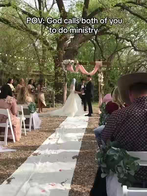 A wide shot captures a wedding ceremony. A bride in a long white gown with a veil stands at an altar, with a groom facing her. Guests are seated, and a long aisle decorated with floral arrangements leads up to the altar. The setting is outdoors, with trees and a natural backdrop.