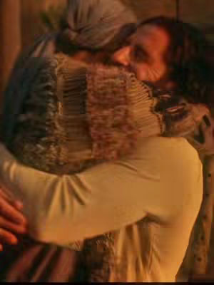 The image shows a warm, close-up embrace between two individuals. One, seen from the back, wears a light-colored garment and a head covering, their profile visible as they lean in. The other person is facing the viewer, with long hair and a partially visible face that is pressed against the other's. The lighting suggests a somewhat dim, interior setting, possibly a room or a building, with a warm color palette dominating.