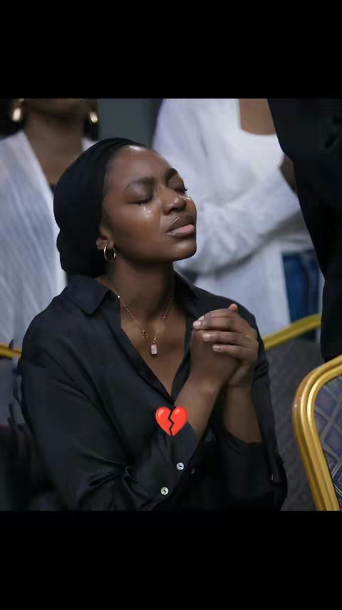 The image shows a woman with visible tears, seemingly in distress. She wears a black head covering and a black button-down shirt. Her hands are clasped together, as if in prayer, and a small, two-part heart graphic is overlaid on her chest. Other people are visible in the blurred background.