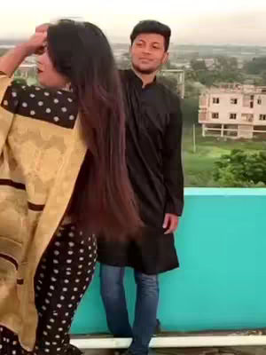 A couple is standing on a rooftop with an urban background. The woman has her back to the camera, and long, dark hair is flowing down. She's wearing a top with white polka dots and a yellow shawl, while the man stands behind her, looking at the camera. They're in a loving pose, suggesting a shared moment. The overall mood is cheerful and relaxed.