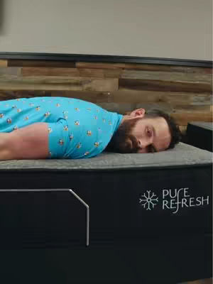 A man is lying face up on a mattress, resting his head against a pillow. He is wearing a blue shirt with a subtle pattern. The mattress is dark and has the words 'Pure Refresh' on the side.