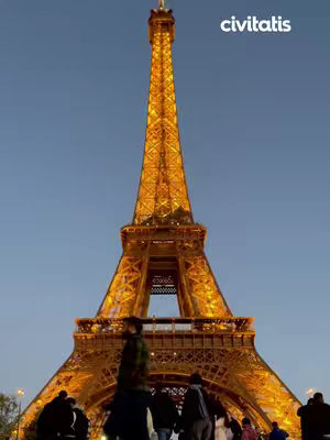 The Eiffel Tower is lit up at night with a blue sky background. A group of people are standing in front of the tower.