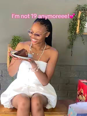 A young woman with dark hair styled in braids is seated outdoors, holding a plate with a slice of chocolate cake. She is wearing sunglasses, a white dress, and jewelry.