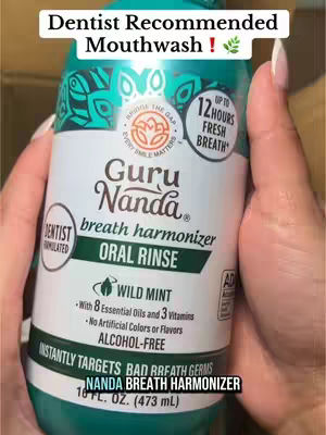 A close-up shot of a person's hand holding a green and white bottle of Guru Nanda mouthwash. The label includes text and logos, with a portion obscured by the person's hand and the background is blurred.