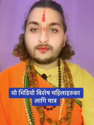 A medium shot features a man with fair skin, dark hair, and a beard set against a solid purple backdrop. He has a red dot on his forehead and wears a yellow top with an orange shawl. He is also wearing multiple layered necklaces with prayer beads. A blue rectangle with white text overlay is positioned at the bottom of the frame.