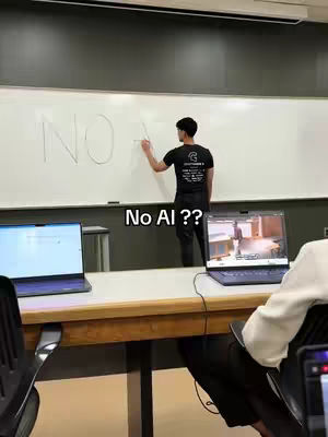 A man is writing on a whiteboard that reads 'NO AI' and gesturing towards it. The room has two laptops open on a table, and someone is seated in the foreground.