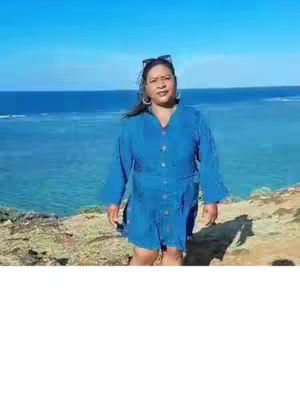 A woman in a blue dress stands on a cliff overlooking the ocean under a clear blue sky. Sunglasses are on her head and the overall image is bright and outdoorsy.