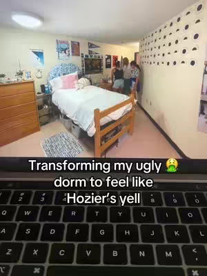 An interior shot of a dorm room, showcasing a bed with a blue headboard and pink accents. Posters and decorations are on the walls. A laptop showing text and keyboard is present at the bottom of the image, with a view of the room behind it.