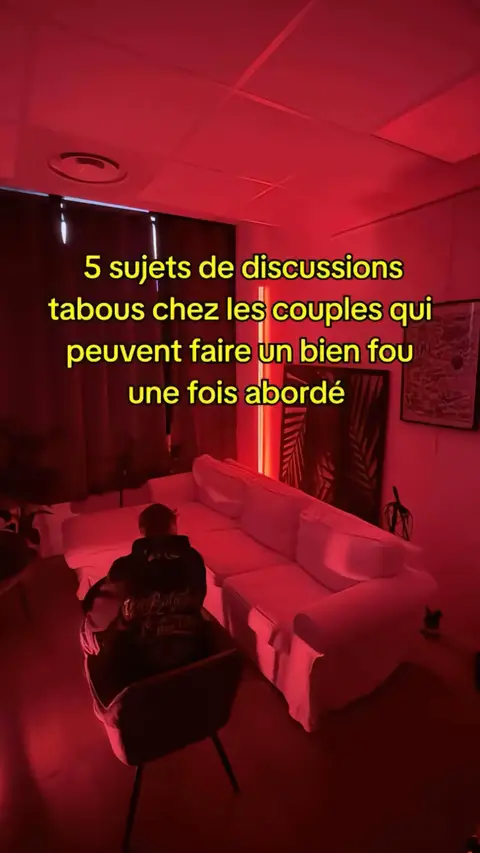 The image shows a dimly lit room with a red ambiance. A white sofa sits on the right, and a person is seated in a chair on the lower left, with their back to the camera. Text overlayed on the center of the picture lists '5 sujets de discussions tabous chez les couples qui peuvent faire un bien fou une fois abordé'.