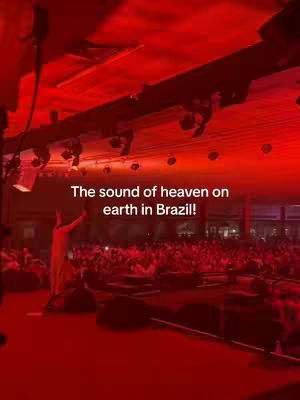 The image shows a large, crowded venue bathed in red light. A figure in a light-colored outfit is on a stage, raising their arms towards the ceiling. The audience is visible in the lower portion of the image. Text overlayed on the image reads: 'The sound of heaven on earth in Brazil!'