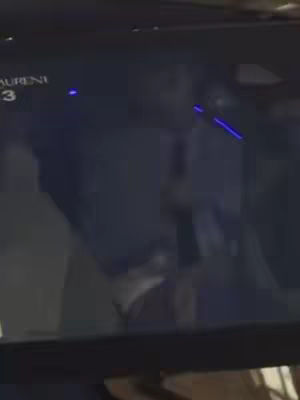 The image is dark, appearing to be a video screen facing an individual in the background. There are some blue light sources on the right of the screen. The subject is hard to distinguish due to the darkness. There are visible details that seem from a car interior.