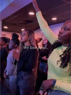 A group of people, primarily women, are gathered in what appears to be a church setting. The lighting is dim with overhead spotlights, and a purple hue tints the background. In the foreground, a woman with a pale green top raises her arm, while others have their hands clasped in prayer. Text overlayed reads 'We love jesus.'
