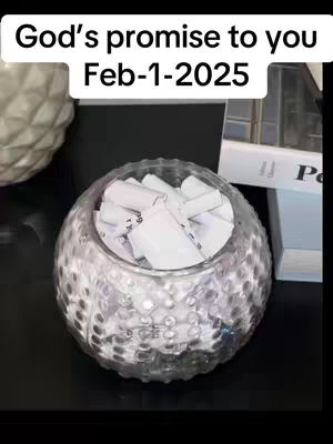 A glass bowl filled with rolled-up papers sits on a dark surface. The bowl has a textured surface. Above it, text indicates a date. The background includes a glimpse of additional objects, suggesting the setting is indoors.