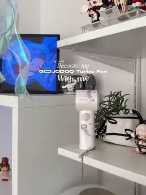 A vertical shot showcasing a white shelf with various items. A desktop fan with decorative stickers is centered. A screen showing an abstract blue graphic is on the left. Other items include figurines, a small bag, and greenery. The color scheme is primarily white with accents of blue and green.