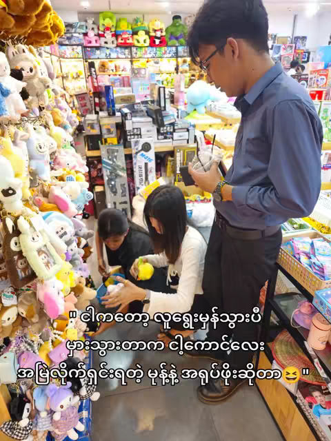 Three people are in a store full of plush toys; A man in a blue shirt is standing on the right side, holding a drink, and appears to be looking at the two people who are near and holding the toys. The store behind them offers a variety of plush toys, and product packages are visible in the background.