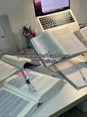 A well-lit, top-down view of a desk featuring an open laptop displaying a screen with text, several open books, a pen, and a notepad. The scene suggests a study environment. The caption 'your goals don't care about your feelings' is overlaid on the image.