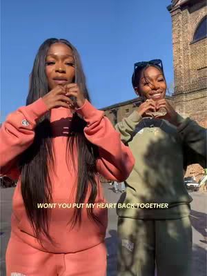Two young women are posing, with each person holding their hands in a heart shape, standing in front of a building.