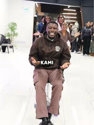 The cover image captures a man in a brown hoodie and pants seated on a stool as a group of people stand behind him. The man is smiling at the camera with his hands open, and there is text overlayed on the image that says "KAMI".