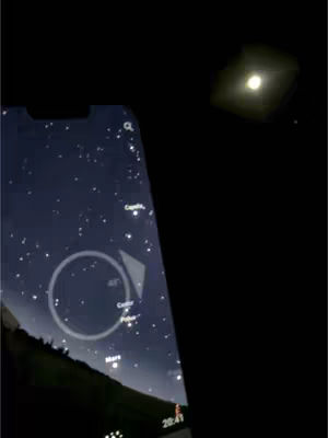 A smartphone screen displaying a star map is held up against a night sky. Above and to the right is a bright light against a black background. The phone display features a circular indicator with the words 'Castor' and 'Pollux' and a line of text 'Mars', while the general background behind it is filled with many tiny stars. The upper right corner has a bright point, likely depicting a planet or star, set against a deep black backdrop.
