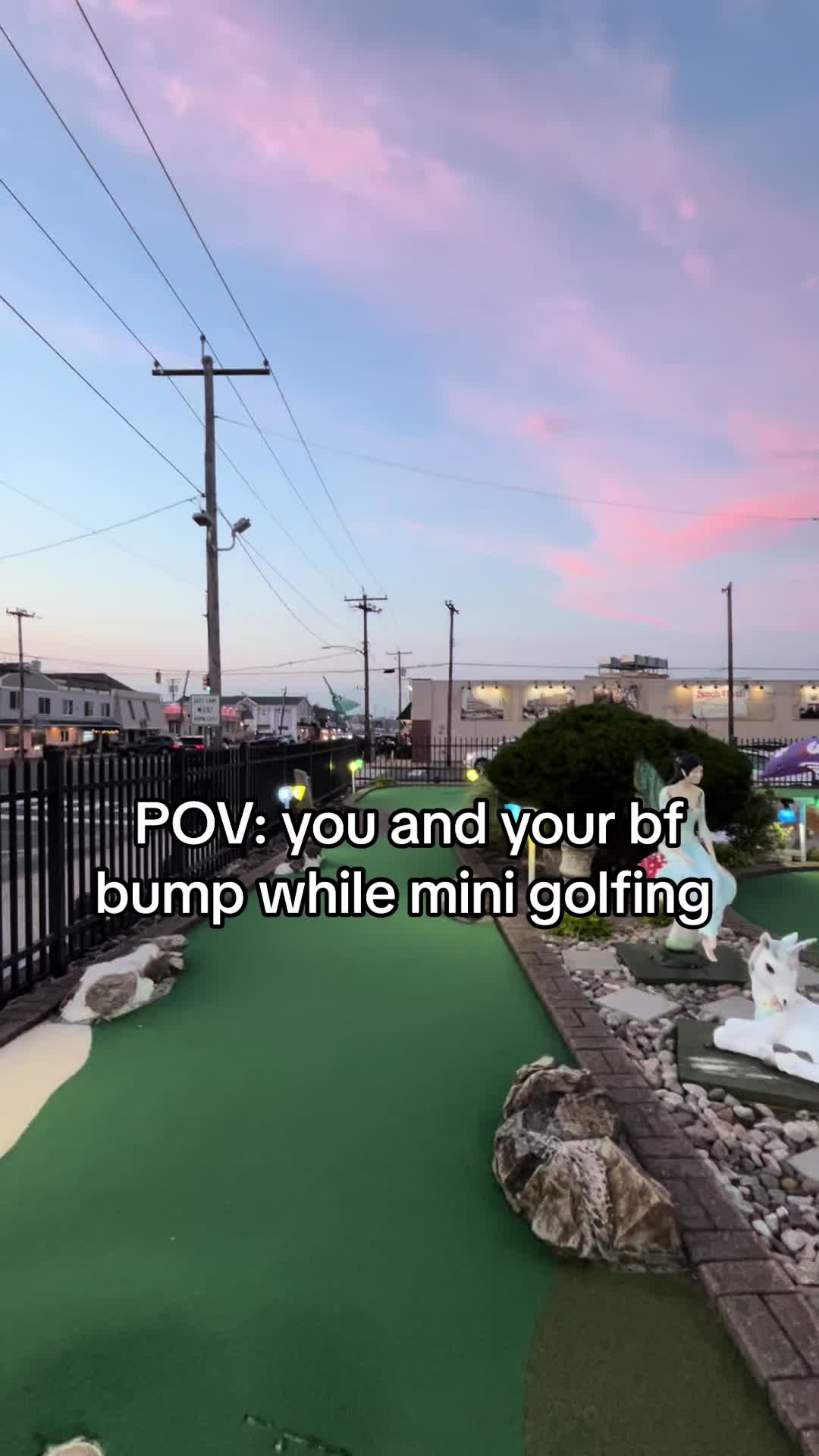 A POV shot at a mini golf course. The sky is filled with pink and blue hues. The scene shows a view of a green, the miniature golf course, and some landscape.