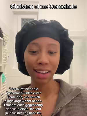 A woman with dark skin is centered in the frame, looking directly at the viewer while speaking. She is wearing a rounded black head covering. Behind her, a white door and a partially visible shelf unit are in the background. The overall color scheme is neutral, with the focus mainly on the woman. There is overlaid text at the top of the screen.