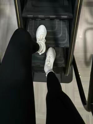 An overhead shot shows a person's feet on a step machine in a gym. Their legs, clad in black leggings, are visible from the knees down. The feet are in white sneakers, positioned on a black step. The background reveals more of the step machine and the gym setting.